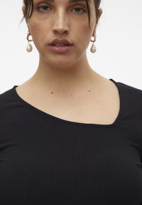 Top negro de canalé con un escote asimétrico único, que presenta una textura suave. La modelo lleva pendientes de perlas colgantes para un toque adicional.