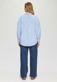 Lichtblauw shirt met lange mouwen en een knoopsluiting, met een rugplooidetail, gecombineerd met donkerblauwe brede jeans en bruine schoenen.