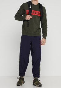 Felpa verde scuro con logo rosso "Calvin Klein", abbinata a pantaloni blu navy con polsini affusolati e pannello laterale verde; zaino nero.