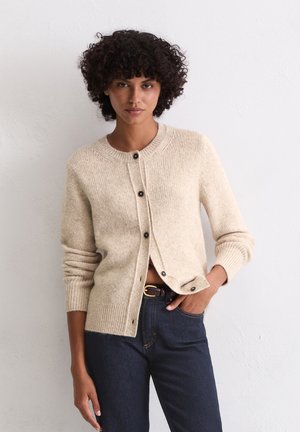 Beige gebreide cardigan met een ronde hals en knoopsluiting. Geribbelde manchetten en taille. Gepaarde met donkerdenim jeans en een bruine riem.