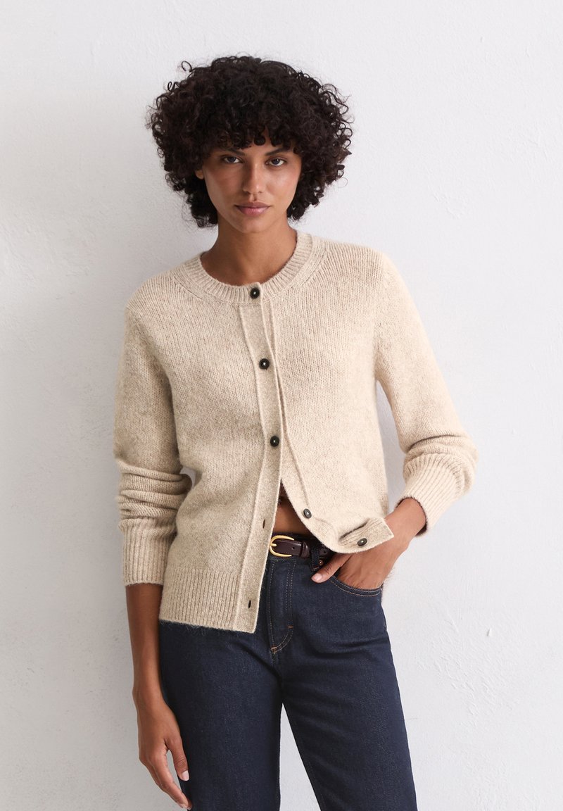 Beige gebreide cardigan met een ronde hals en knoopsluiting. Geribbelde manchetten en taille. Gepaarde met donkerdenim jeans en een bruine riem.