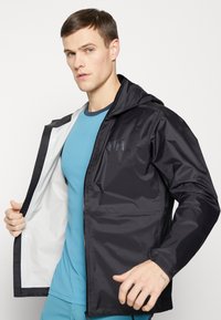 Chaqueta negra impermeable con capucha, dos bolsillos frontales y un forro blanco. Lleva puesta sobre una camiseta azul ajustada. Textura suave, diseño ligero.