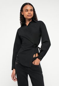 Μαύρο πουκάμισο τύπου wrap από βαμβακερό μείγμα με μακριά μανίκια, γιακά, λεπτομέρεια δέσιμου στο πλάι και κεντημένο λογότυπο. Συνδυασμένο με μαύρα τζιν, λεία υφή.