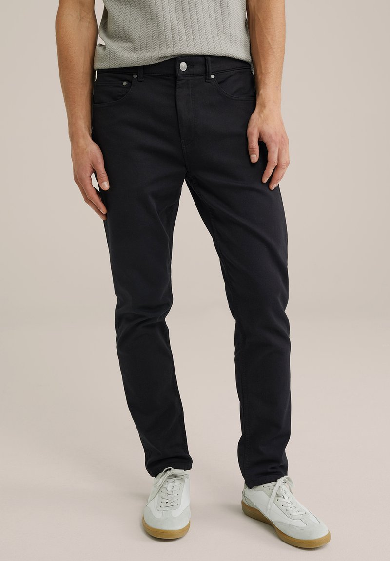 Sötétkék, slim-fit farmernadrág, pamutból készült, közepes derékkal, öt zsebbel és finom varrási részletekkel. Világos sportcipővel párosítva.