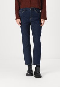 Tumši zila džinsu auduma bikses ar taisnu griezumu, piecu kabatu dizainu un kontrastējošu šuvēm, apvienotas ar melniem slip-on apaviem.