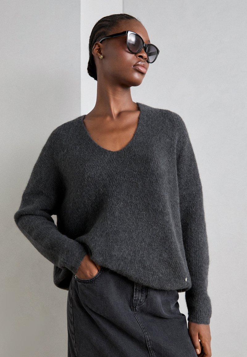Grauer, übergroßer Pullover mit V-Ausschnitt, langen Ärmeln und weicher Textur. Kombiniert mit einem dunklen Rock; das Model trägt schwarze Sonnenbrillen.