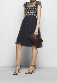 Robe en tulle noire avec broderies florales et manches volantes ; dotée d'un corsage ajusté et d'une jupe évasée mi-genoux. Tenant une pochette en osier marron.