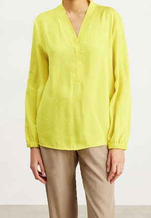 Personne portant un chemisier jaune vif à manches longues avec un col en V et un pantalon beige, visible du cou jusqu'au milieu de la cuisse.