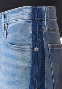 Blå denimjeans med en tvåfärgad design, där ett ljusare och ett mörkare blått material kombineras. Har standardfickor och gula sömnadsdetaljer.