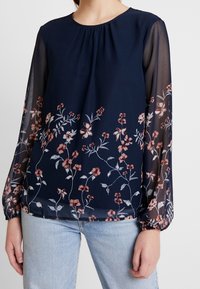 Navy blue paita, jossa on pitkät läpikuultavat hihat, ja helmassa on kukka-ompeleet vaaleanpunaisissa ja valkoisissa sävyissä. Pyöreä pääntie, rypytetty yksityiskohta.