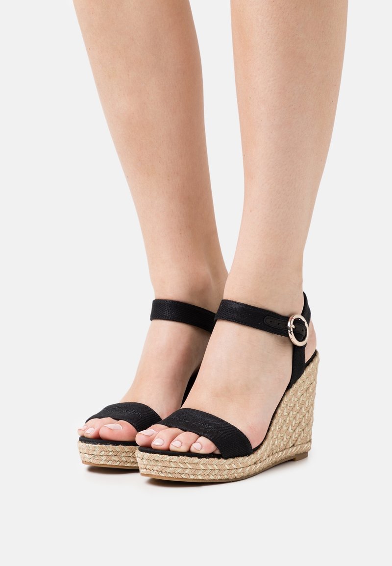 Sandales compensées noires avec une texture tissée d'espadrille, dotées d'une large bride sur les orteils et d'une bride de cheville réglable avec une boucle argentée.