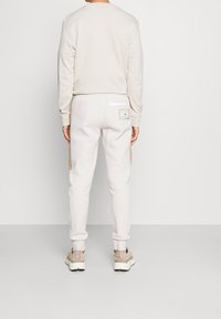 Lys beige sweatshirt med rund hals og matchende sweatpants. Bukserne har en baglomme med en hvid lap og brune detaljer.