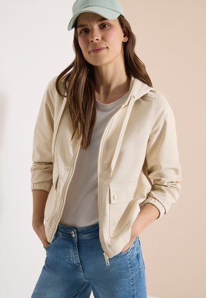 Kobieta ubrana w jasnobeżową kurtkę na zamek z kieszeniami, białą koszulkę, niebieskie dżinsy i jasnoniebieską czapkę z daszkiem, ręce w kieszeniach.