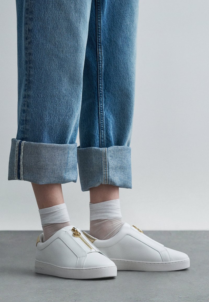 Baskets blanches à enfiler avec un détail zippé, tige en cuir texturé et semelle en caoutchouc blanche, portées avec un pantalon en denim bleu retroussé.