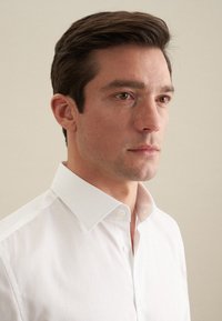 Witte button-up shirt met een spitse kraag, gemaakt van soepel katoen en met een getailleerde pasvorm. Het model heeft een neutrale uitdrukking.