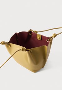 Otvorena kožna torba boje gorušice s uvijenim naramenicama i glatkom bordo unutrašnjom oblogom na bijeloj pozadini.