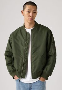 Ung mann med kort hår iført olivengrønn bomberjakke, hvit t-skjorte og lyseblå jeans, hendene i lommene, nøytral bakgrunn.