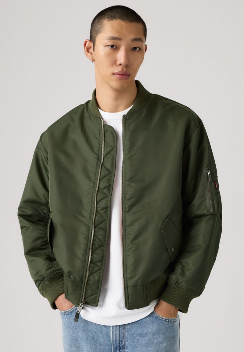 Ung mann med kort hår iført olivengrønn bomberjakke, hvit t-skjorte og lyseblå jeans, hendene i lommene, nøytral bakgrunn.