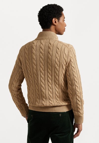 Suéter de malha cabos na cor bege, com colarinho alto, apresentando padrões torcidos verticais e punhos e bainha canelados. Usado sobre calças de veludo cotelê verde-escuro.