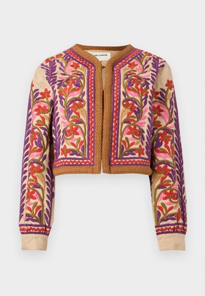 Femme portant une veste courte brodée sur une blouse blanche, un pantalon noir à jambes larges, des talons blancs, et tenant un petit sac à main rond orange.