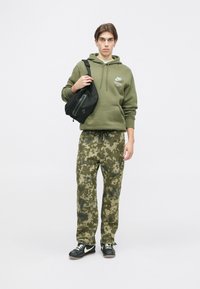 Oliiviroheline kapuutsiga dressipluus logoga, koos heledate kamuflaažimustriga dressipükstega ja mustade spordijalatsitega. Õlal on peen kott.