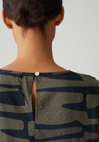 Green and navy patterned top with a scoop neckline, featuring a back button closure and textured fabric.
