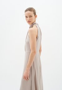 Ärmelloses beiges Kleid mit einem drapierten Kragen, weicher Textur, bodenlanger Silhouette und einem seitlichen Schlitz.
