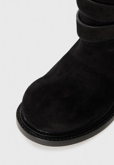 Czarna zamszowa cholewka buta z podwójnym paskiem oraz grubą szytą podeszwą na białym tle.