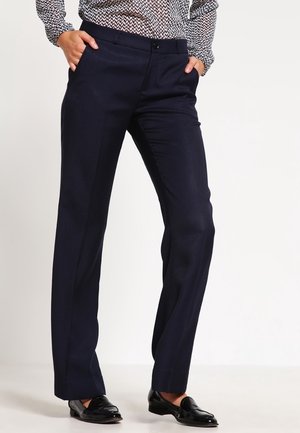 Woman wearing black straight-leg trousers with hands in pockets, paired with black loafers and a patterned long-sleeve top.