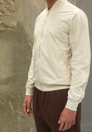 Homme portant une veste zippée Nike beige clair et un pantalon marron foncé, debout contre un mur texturé.