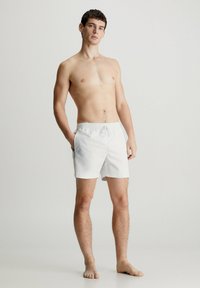 Costumi da bagno bianchi con vita elastica e cordoncino, dotati di tasche laterali. Il tessuto appare leggero e liscio.