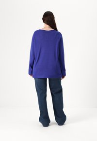 Pulover tricotat violet cu decolteu larg și mâneci lungi, combinat cu pantaloni din denim închis și largi, văzut din spate pe un fundal simplu.