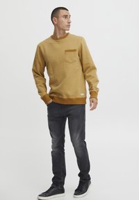 Sudadera marrón de jersey con cuello redondo y un bolsillo en el pecho, combinada con jeans gris oscuro y zapatillas negras con suelas blancas.