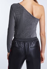 Asymmetrise metallinen bodysuit tummanharmaana, jossa on yksi pitkä hiha, yhdistettynä mustiin keinonahkahaylareihin, joissa on nyörikiinnitys vyötäröllä.