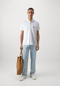 Polo shirt bianco con un piccolo logo scuro, jeans azzurri chiari e sneakers bianche. Una borsa a tracolla marrone è tenuta nella mano sinistra.