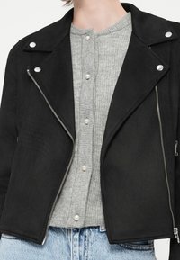 Giacca nera in ecopelle scamosciata con hardware argentato, chiusura con zip e bottoni a scatto. Indossata sopra un top grigio a coste con bottoni in perla.