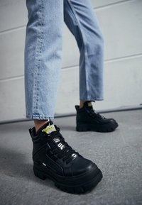 Baskets montantes noires avec un extérieur en cuir texturé, une semelle épaisse et des accents superposés, assorties à un jean clair retroussé.