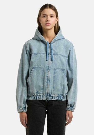 Ung kvinne iført lyseblå denimjakke med glidelås, hette med snorer og elastiske mansjetter, kombinert med svarte bukser, står stille.