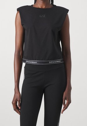 Femme portant un haut noir sans manches avec des épaules rembourrées et un pantalon noir assorti avec des bandes logo EA7 Emporio Armani.