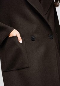 Abrigo de lana marrón con cierre de botones de doble fila, solapas con muesca y un bolsillo lateral, con una textura suave y un ajuste entallado.