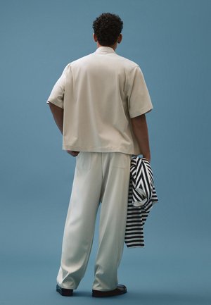 Man with curly hair stands facing away, wearing beige short-sleeve shirt and pants, holding a black-and-white striped garment against a blue background.