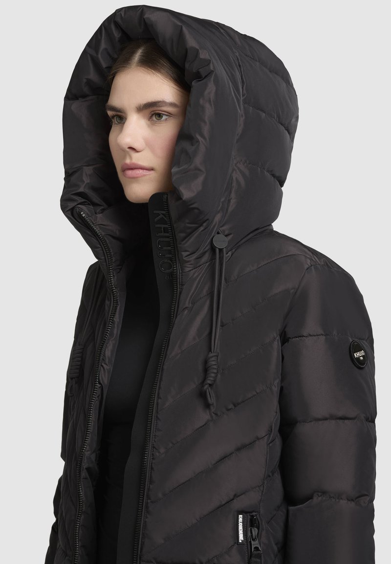 Schwarze wattierte Jacke mit einer breiten, gefütterten Kapuze, gestepptem Material, Frontreißverschluss und Zugband-Akzenten. Mit Logo auf dem Ärmel.