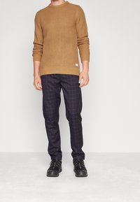 Stickad tröja i senapsgul med ett texturerat mönster, kombinerad med navy-rutiga byxor. Svarta skor med en robust sula synlig.