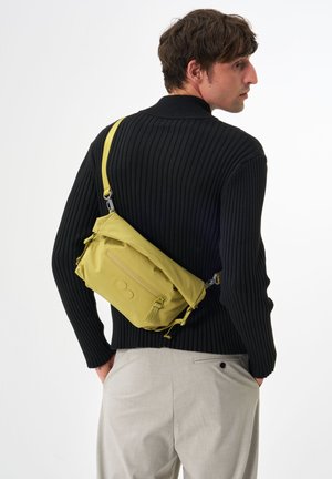 Gelbe Nylon Crossbody-Tasche mit einer Reißverschlusstasche vorne, verstellbarem Riemen und Kordelzug-Detail, getragen über einem schwarzen gerippten Pullover.