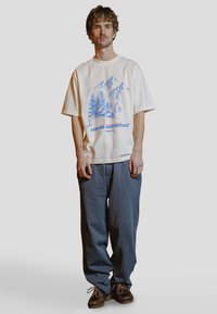 Ung mann med krøllete hår står, kledd i en hvit T-skjorte med blått fjellmotiv, løstsittende grå bukser og brune snøresko.