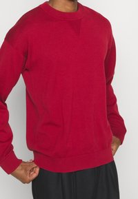 Roter Baumwoll-Sweater mit Rundhalsausschnitt und Rippstrickkragen sowie -abschlüssen, mit strukturiertem Detail auf der Brust und lässiger Passform.