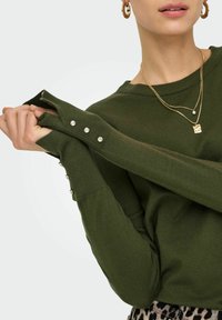 Olivgrüner Strickpullover mit gerippten Bündchen, der Perlmuttknopf-Akzente an den Ärmeln aufweist. Der Stoff wirkt weich und hat einen lässigen Schnitt.