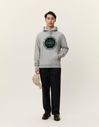 Γκρι φούτερ με κουκούλα που διαθέτει μπροστινή γραφική παράσταση με το κείμενο "LES DEUX COPENHAGEN" σε σκούρο πράσινο. Συνδυάζεται με μαύρο παντελόνι και μπεζ καπέλο.