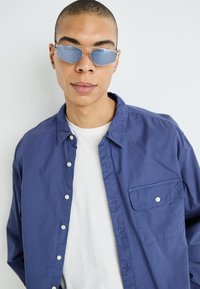 Camisa de botones azul marino con un bolsillo, llevada sobre una camiseta blanca de cuello redondo. Gafas de sol tipo aviador plateadas con lentes azules. Textura de tela suave.