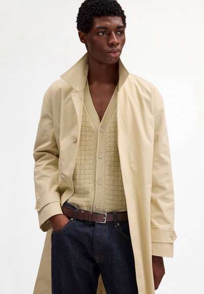 Jeune homme portant un trench-coat beige, un cardigan beige à boutons texturé, une ceinture marron et un jean bleu foncé, debout avec une main dans la poche.